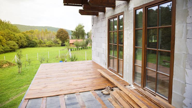 Blick auf eine teilweise fertiggestellte Holzterrasse mit Baumaterialien und Fensterfront eines Hauses im Grünen.